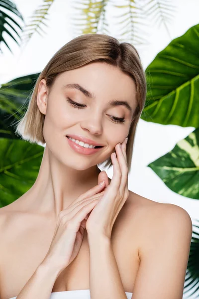 Усміхнена молода жінка з ідеальною шкірою біля зеленого пальмового листя на білому — стокове фото