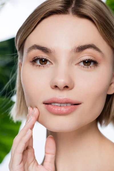 Close up view of blonde young woman with perfect skin looking at camera near green palm leaves on white — Stock Photo