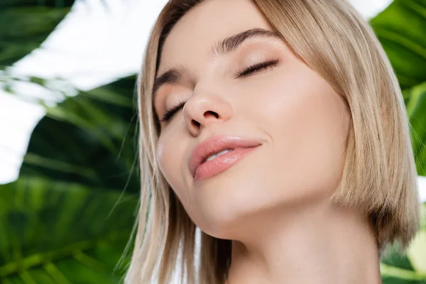 Primer plano de mujer joven con la piel perfecta con los ojos cerrados cerca de hojas de palma verde sobre blanco - foto de stock