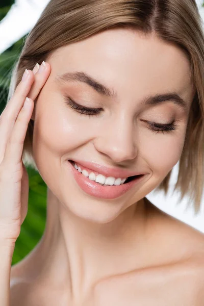 Gros plan de jeune femme heureuse avec une peau parfaite regardant vers le bas près de feuilles de palmier vert sur blanc — Photo de stock