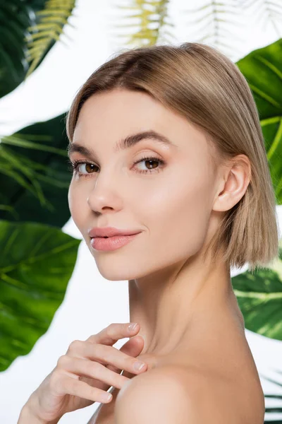 Blonde woman with perfect skin looking at camera near palm leaves on white — Stock Photo
