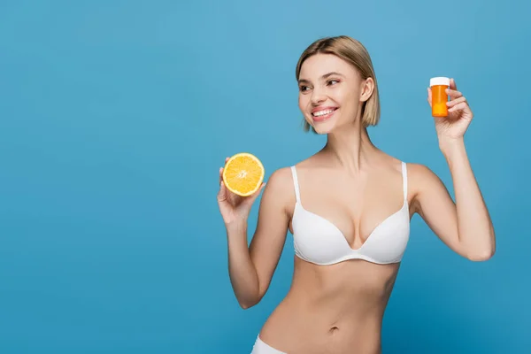 Jovem alegre em lingerie branca segurando garrafa com vitaminas e laranja metade isolado em azul — Fotografia de Stock
