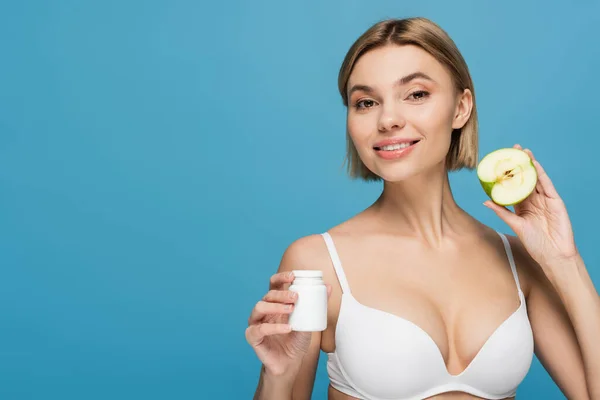 Heureuse jeune femme en lingerie blanche tenant bouteille avec vitamines et pomme à moitié isolé sur bleu — Photo de stock