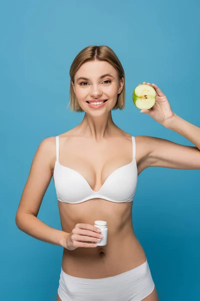 Cheerful woman in white lingerie holding bottle with vitamins and apple half isolated on blue — Stock Photo