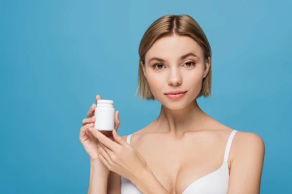 Mujer joven rubia en sujetador blanco sosteniendo botella con vitaminas aisladas en azul - foto de stock