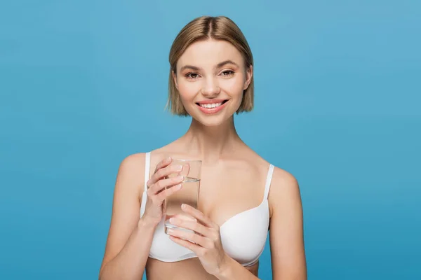 Happy young woman in white bra holding glass of water isolated on blue — Stock Photo