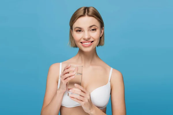 Feliz joven en sujetador blanco sosteniendo vaso de agua aislado en azul - foto de stock