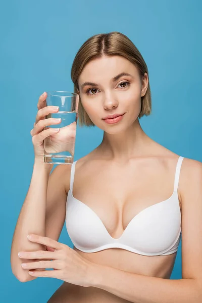Блондинка молода жінка в білому бюстгальтері тримає склянку води ізольовано на синьому — стокове фото
