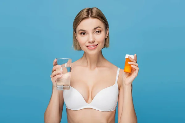 Joyful young woman in white bra holding bottle with vitamins and glass of water isolated on blue — Stock Photo