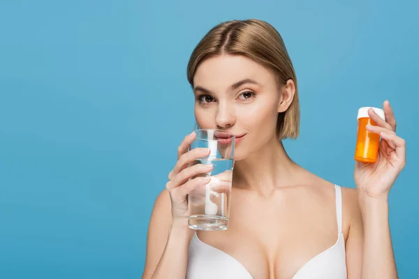 Junge Frau in weißem BH hält Flasche mit Vitaminen und Glas Wasser isoliert auf blau — Stockfoto