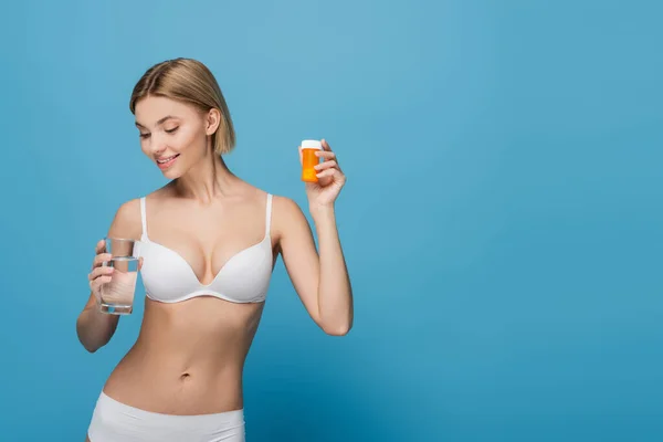 Sonriente joven en sujetador blanco sosteniendo botella con vitaminas y vaso de agua aislado en azul - foto de stock