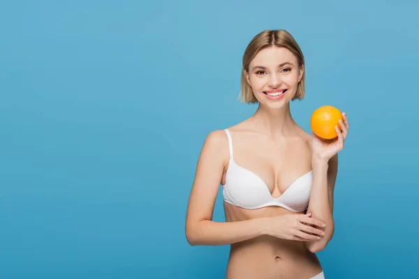 Sorridente giovane donna in reggiseno bianco tenendo maturo arancione isolato su blu — Foto stock