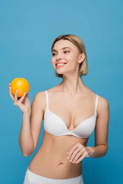 Весела молода жінка в білому бюстгальтері тримає стиглий апельсин ізольовано на синьому — стокове фото