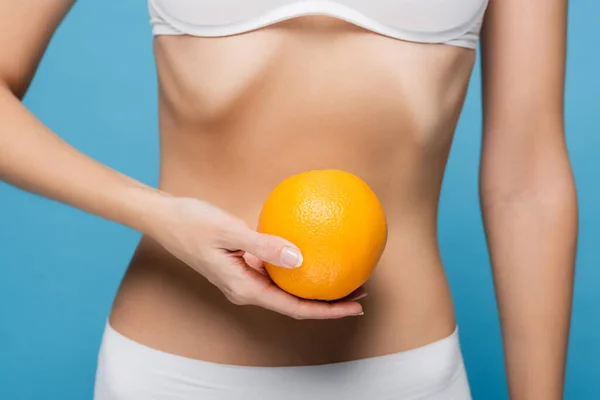 Обрізаний вид на молоду жінку в білому бюстгальтері, що тримає стиглий апельсин ізольований на синьому — стокове фото