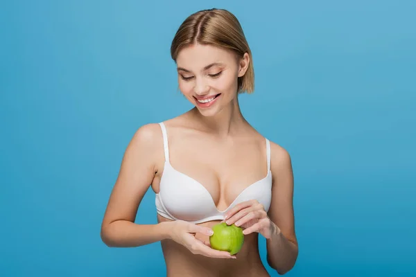 Весела молода жінка в білому бюстгальтері тримає стигле зелене яблуко ізольовано на синьому — стокове фото