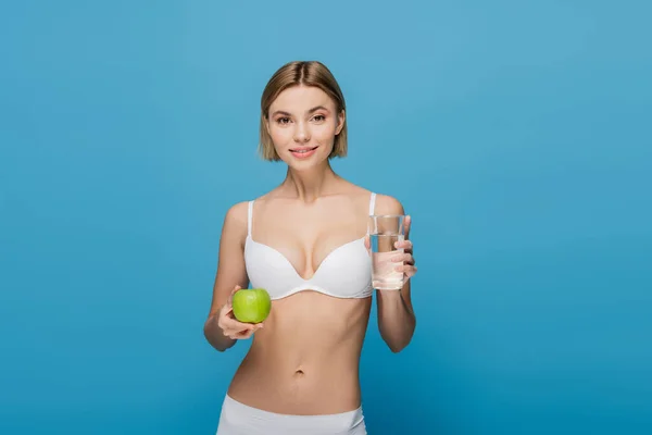 Усміхнена молода жінка в білому бюстгальтері тримає зелене яблуко і склянку води ізольовані на синьому — стокове фото
