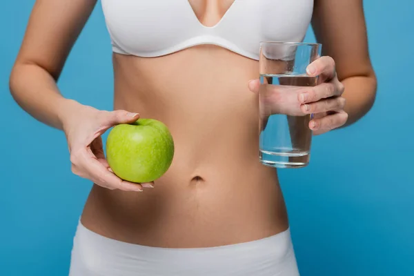 Abgeschnittene Ansicht einer jungen Frau in weißer Unterwäsche mit grünem Apfel und Glas Wasser isoliert auf blauem Grund — Stockfoto