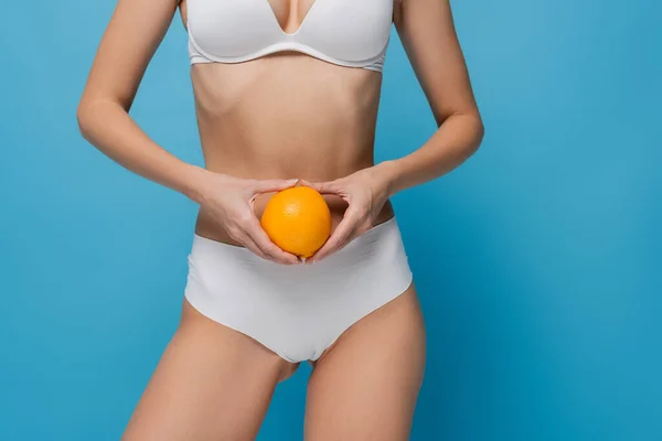 Cropped view of young woman in white lingerie holding ripe orange isolated on blue — Stock Photo