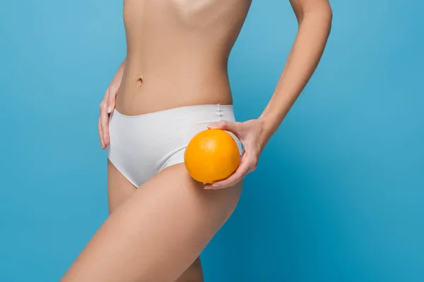 Ausgeschnittene Ansicht einer schlanken jungen Frau in weißen Dessous, die ganze Orange isoliert auf Blau hält — Stockfoto