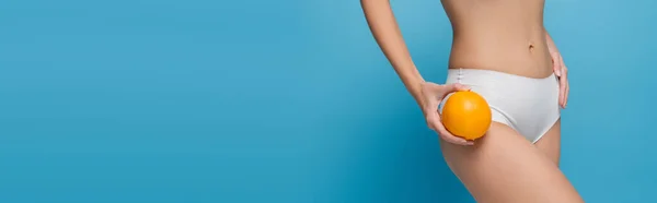 Cropped view of slim young woman in white lingerie holding ripe orange isolated on blue, banner — Stock Photo