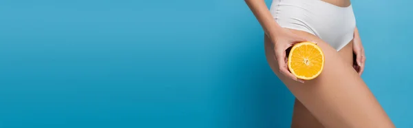 Cropped view of slim woman in white lingerie holding half of orange isolated on blue, banner — Stock Photo