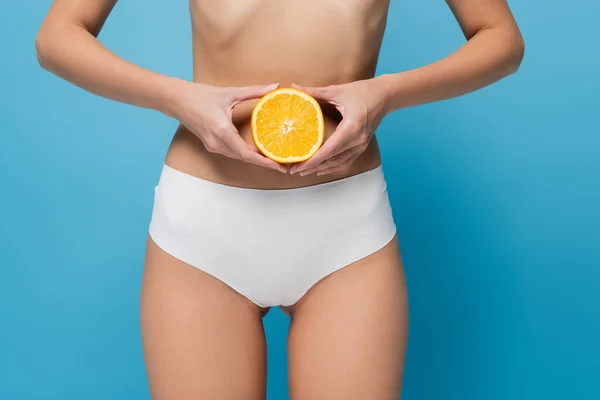 Partial view of young slim woman in white panties holding half of orange isolated on blue — Stock Photo