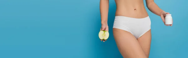 Cropped view of slim woman in white panties holding half of apple and bottle with pills isolated on blue, banner — Stock Photo