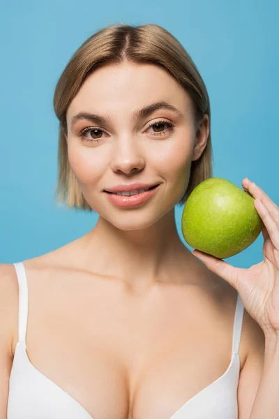 Blonde jeune femme en soutien-gorge tenant pomme verte et souriant isolé sur bleu — Photo de stock