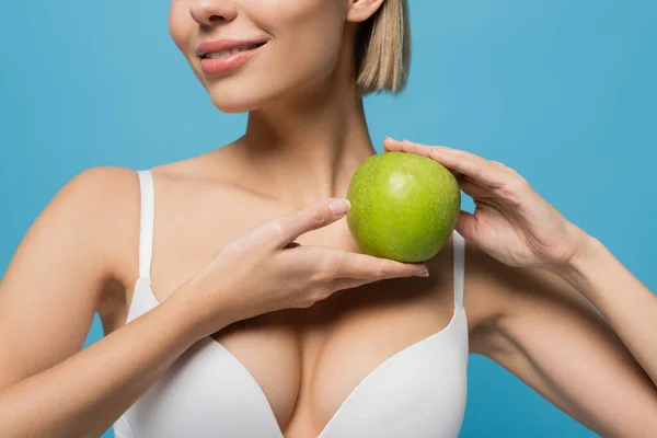 Recortado vista de la joven rubia en sujetador blanco sosteniendo manzana verde y sonriendo aislado en azul - foto de stock