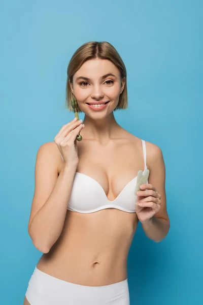 Cheerful young woman in white bra holding jade scrapper and roller isolated on blue — Stock Photo