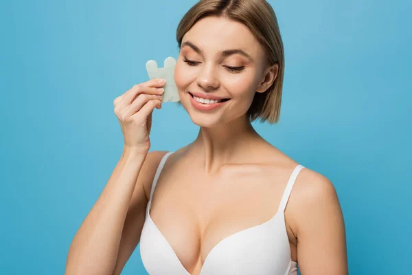 Cheerful young woman in bra massaging face with jade scrapper isolated on blue — Stock Photo