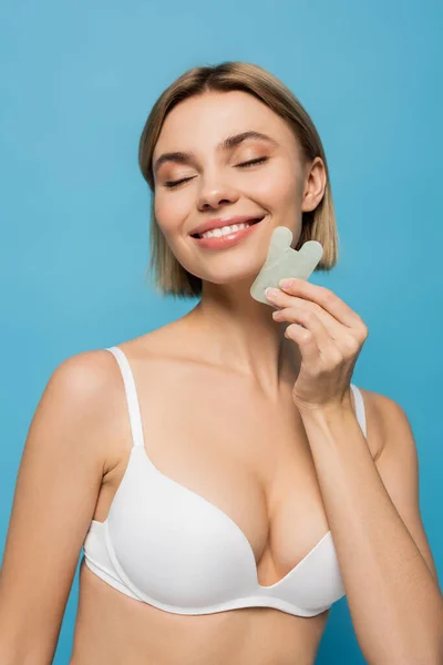 Fröhliche junge Frau im BH massiert Gesicht mit Jade-Werkzeug isoliert auf blau — Stockfoto