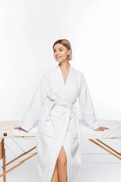Jovem mulher feliz em roupão de banho perto da mesa de massagem isolada no branco — Fotografia de Stock
