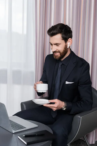 Cheerful bearded man in earphone holding cup and looking at laptop — Stock Photo