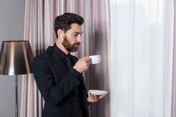 Hombre barbudo en auricular sosteniendo taza y platillo en la habitación de hotel - foto de stock