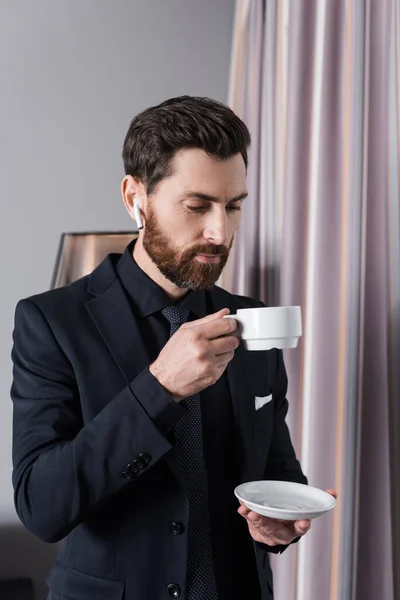 Bearded businessman in earphone holding cup and saucer in hotel room — Stock Photo