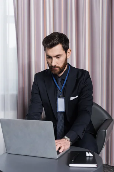 Hombre de negocios barbudo con placa de identificación utilizando el ordenador portátil en la habitación de hotel - foto de stock