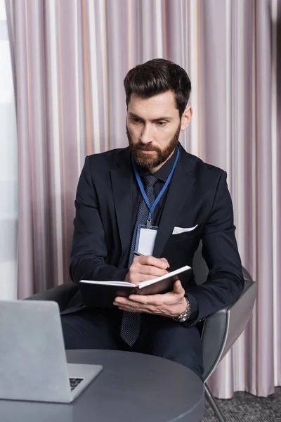 Uomo d'affari barbuto con id badge scrittura nel taccuino e guardando il computer portatile in camera d'albergo — Foto stock