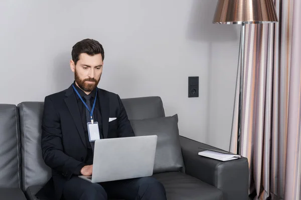 Bearded businessman with id badge using laptop while sitting on leather couch in hotel room — Stock Photo