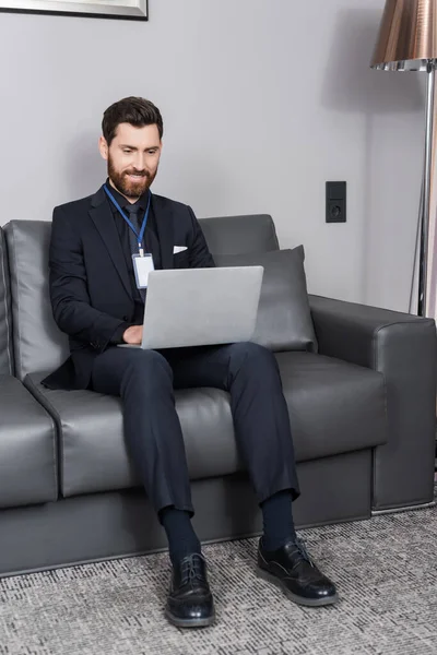Alegre hombre de negocios con placa de identificación utilizando el ordenador portátil mientras está sentado en el sofá de cuero en la habitación de hotel - foto de stock