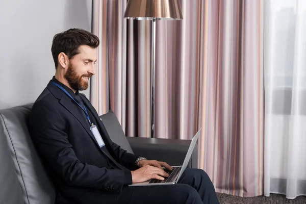 Joyful businessman with id badge using laptop while sitting on leather couch in hotel room — Stock Photo