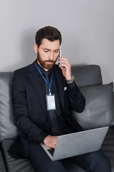 Bearded businessman with id badge talking on smartphone and using laptop in hotel room — Stock Photo