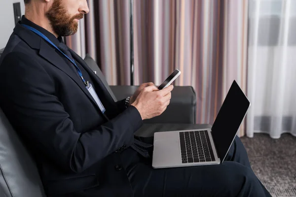 Vista ritagliata di uomo d'affari barbuto con badge id utilizzando smartphone vicino laptop in camera d'albergo — Foto stock