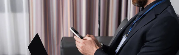 Vista recortada de hombre de negocios con placa de identificación utilizando el teléfono inteligente cerca de la computadora portátil en la habitación del hotel, pancarta - foto de stock