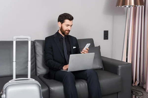 Alegre hombre de negocios con teléfono inteligente cerca de la computadora portátil y el equipaje en la habitación del hotel - foto de stock