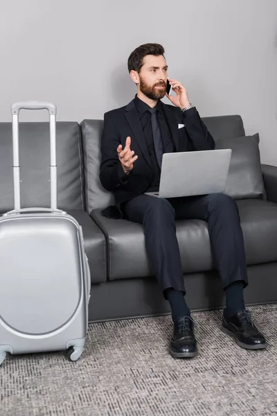 Homem de negócios barbudo falando em smartphone perto de laptop e bagagem no quarto de hotel — Fotografia de Stock