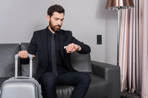 Hombre de negocios barbudo comprobando el tiempo en reloj de pulsera y sentado cerca de equipaje en la habitación del hotel - foto de stock