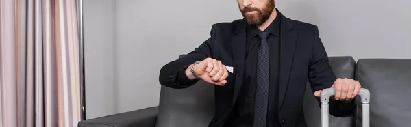 Vista recortada del hombre de negocios barbudo comprobando el tiempo en reloj de pulsera y sentado cerca de equipaje en la habitación del hotel, pancarta - foto de stock