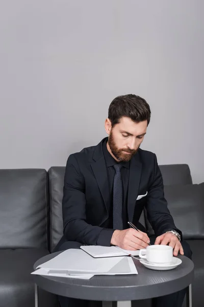 Homem barbudo em terno escrevendo no notebook perto de documentos no quarto de hotel — Fotografia de Stock