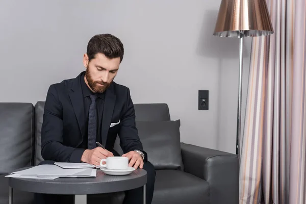 Homme d'affaires barbu en costume écrit sur ordinateur portable près des documents et tasse de café dans la chambre d'hôtel — Photo de stock